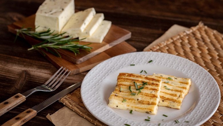 Grilled cheese on a plate decorated with herbs, next to it a wooden board with cheese slices and herbs.