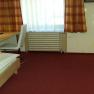 A simply furnished hotel room with a single bed, desk and chair. Red carpet and checkered curtains.