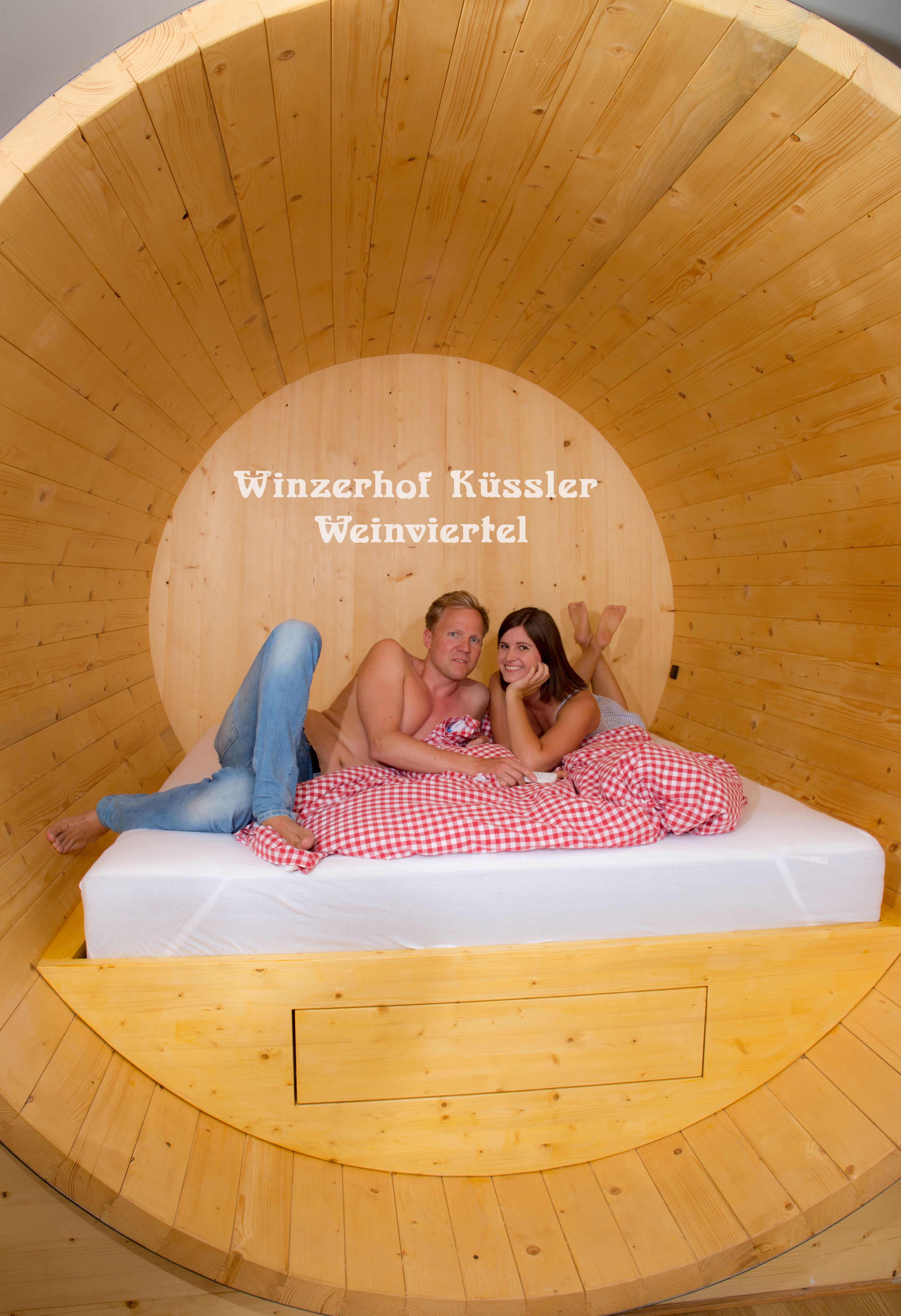 A couple lies in a large wooden barrel bed with red and white checkered bedding. The words 'Winzerhof Küssler Weinviertel' are in the background.