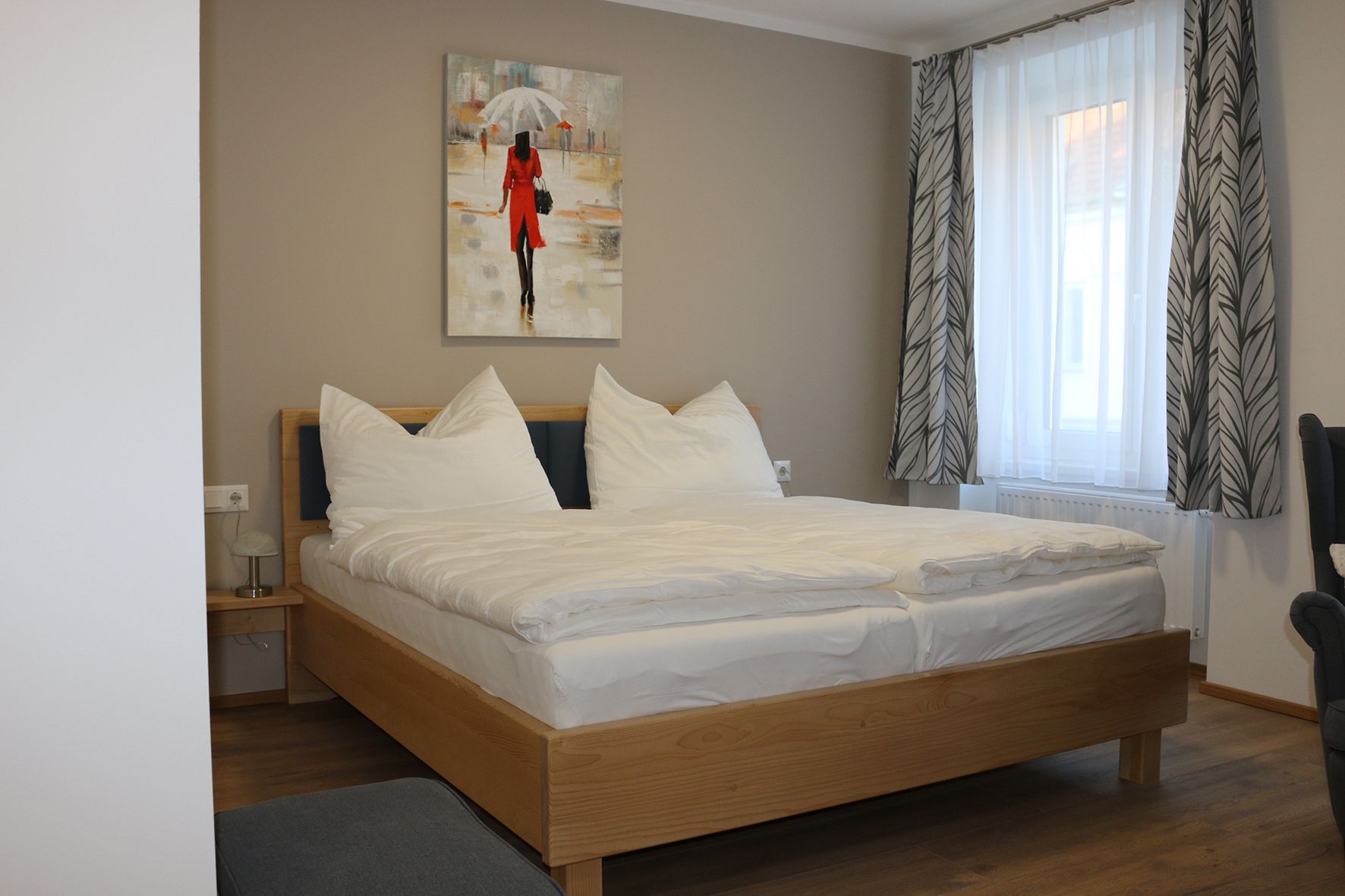 Wooden double bed with white bed linen. Behind it a window with curtains and a painting.