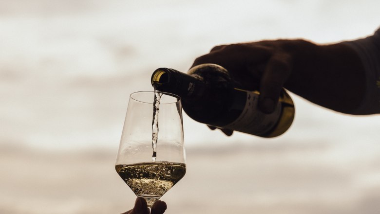 A hand pours white wine into a glass outside, in front of a cloudy sky.