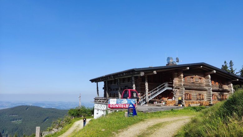 A wooden mountain hut with a red vehicle in front of it, surrounded by green meadows and a clear blue sky.