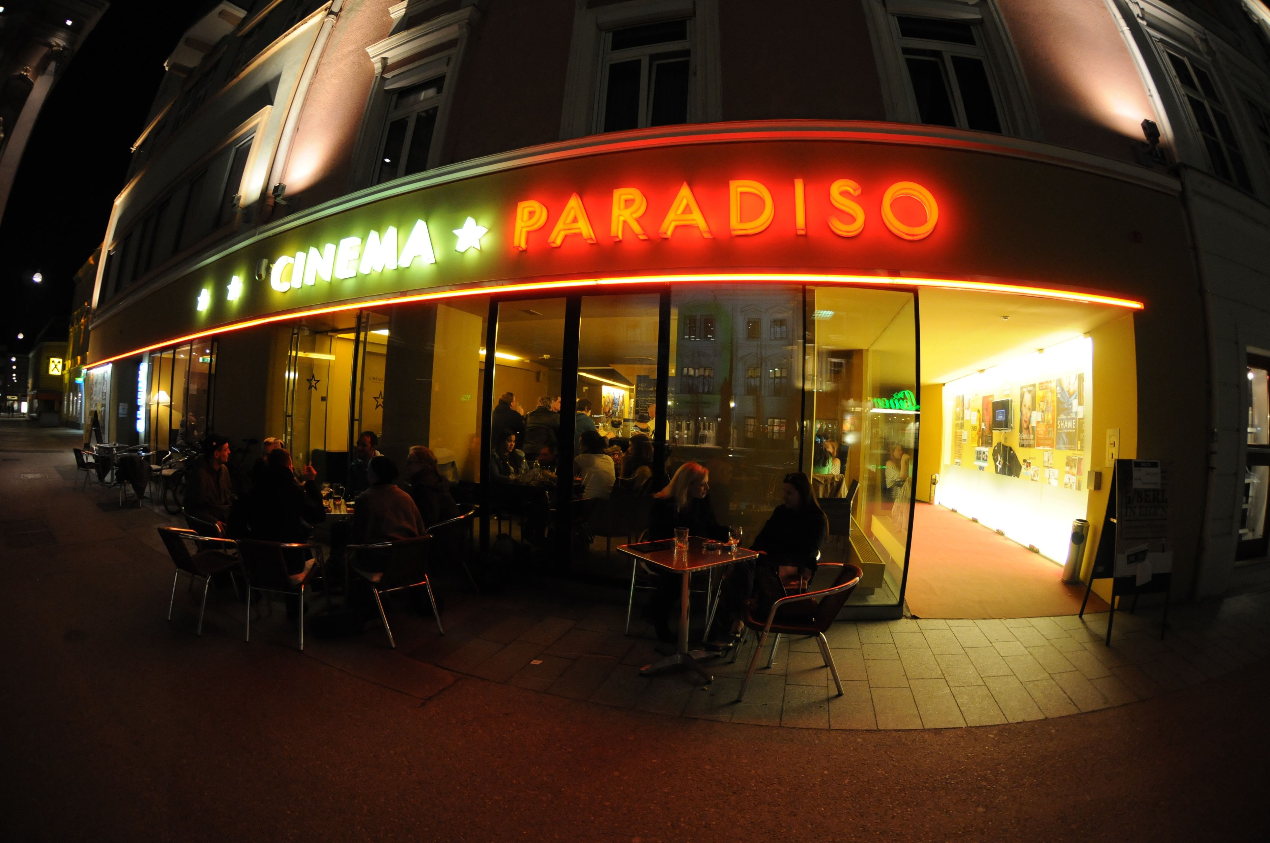 Exterior view of a movie theater called Cinema Paradiso at night with illuminated entrance and people at tables.
