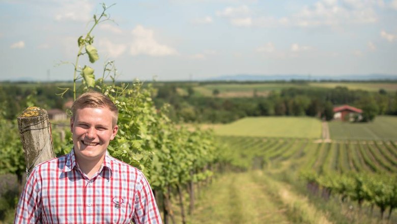 A smiling man stands in a vineyard with a sweeping view over the landscape.