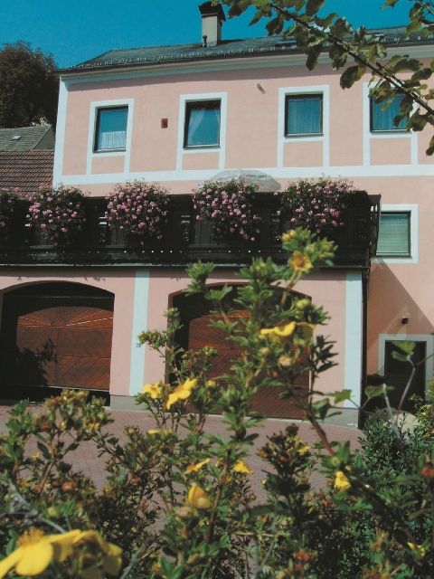 A two-storey pink building with flowering plants on the balcony and yellow flowers in the foreground.