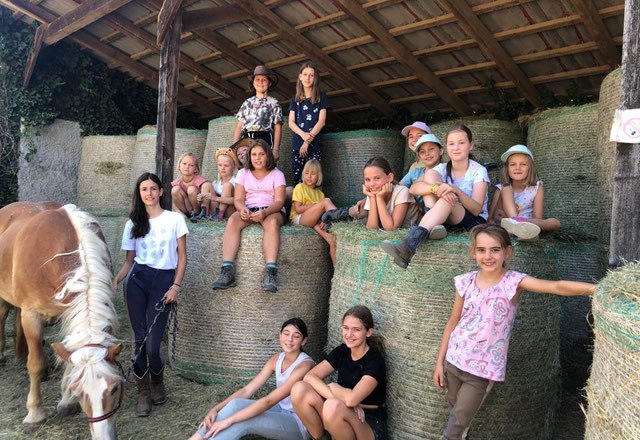 A group of children are sitting on hay bales in a stable, with a horse standing next to them. The children are smiling and wearing casual clothes.