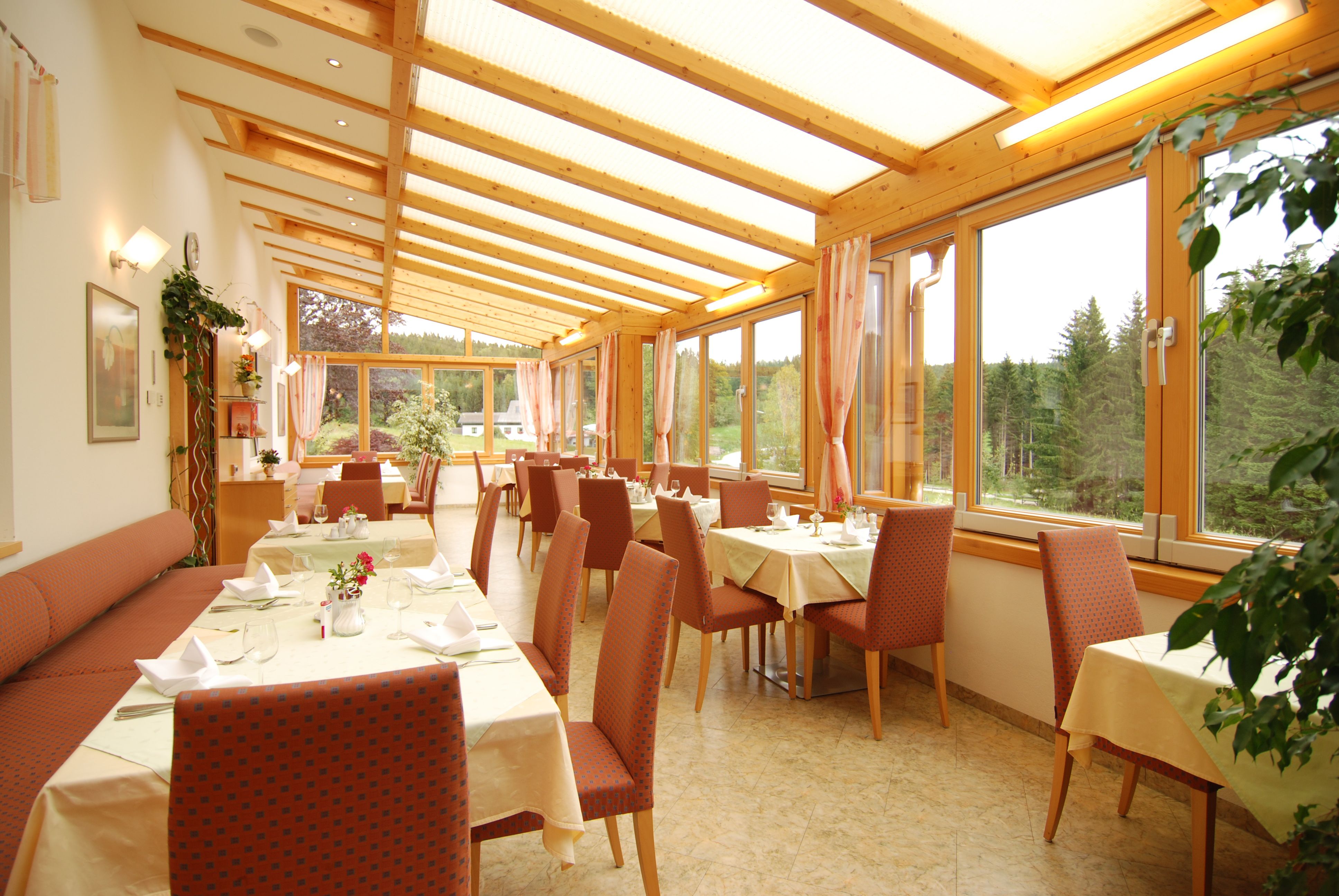A bright conservatory of a restaurant with set tables and a view of a forest.