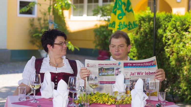 A man and a woman are sitting at an outdoor table set with glasses and cutlery. The man is reading a newspaper.