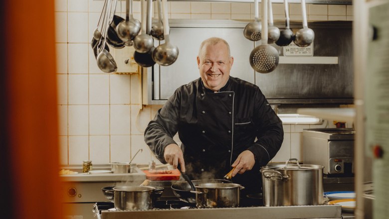 A chef in a black uniform stands smiling in a kitchen and cooks at a stove with several pots.