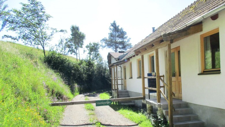 White building with a barrier with the sign "Tavern" next to a gravel path and a green meadow with trees.