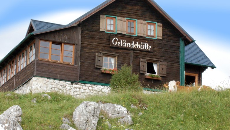 A mountain hut with the inscription 'Geländehütte' on a meadow with rocks and a cow in the foreground.