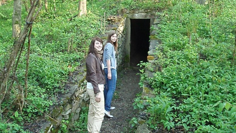 Two people are standing in front of an old ice cellar in the forest.