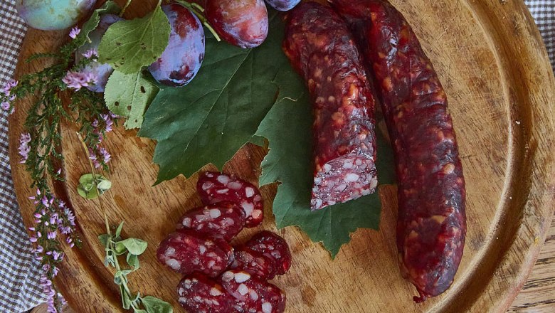 Kaminwurzn and plums on a wooden board decorated with green leaves and herbs.