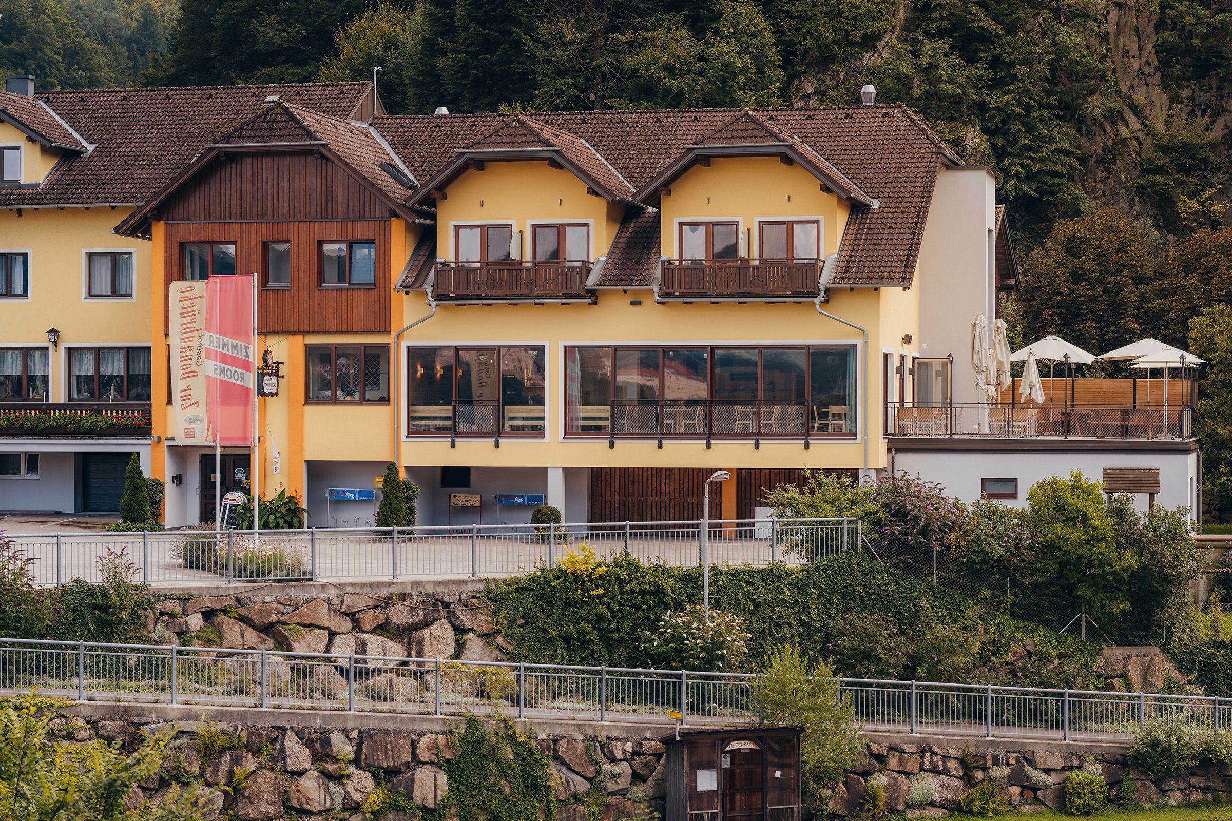 Yellow building with terrace and signs, surrounded by trees.
