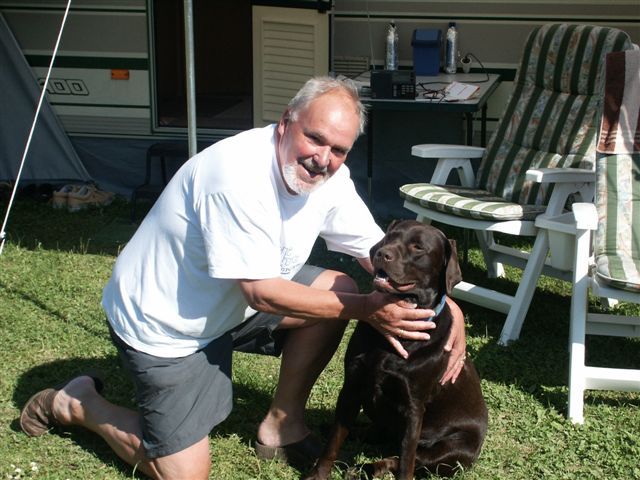 A man kneels next to a brown dog on a meadow in front of a caravan.