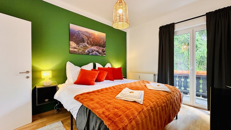 Cozy bedroom with green painted wall, double bed with orange blankets and pillows, lamp and balcony.