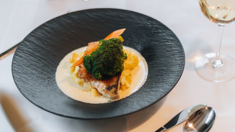 An elegantly presented dish on a black plate with vegetables and sauce, next to a glass of white wine on a laid table.