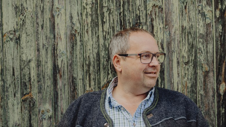 Man with glasses in front of a weathered wooden wall.