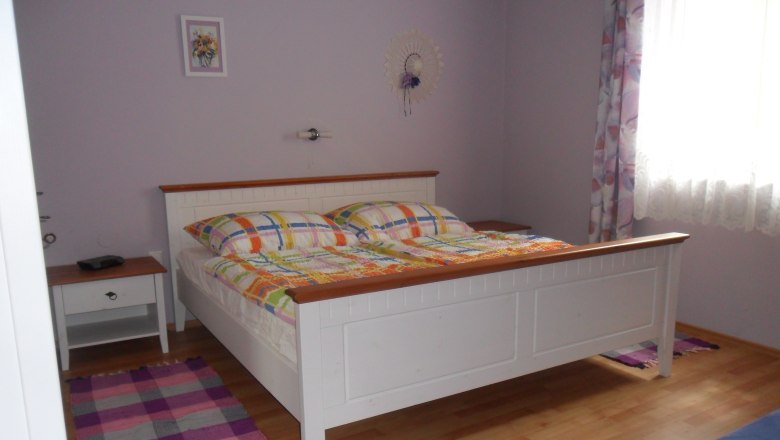 Bedroom, © Fam. Faber A bedroom with a white bed, colorful comforters, a bedside table and a window with curtains.