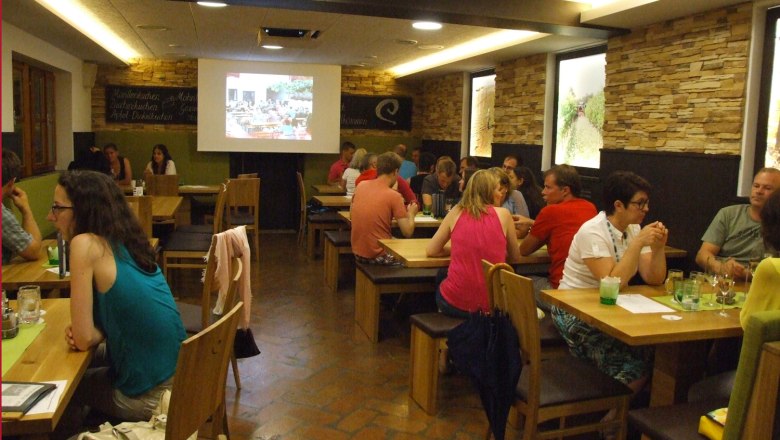People sit at wooden tables in a cozy room while a presentation is shown on a screen.