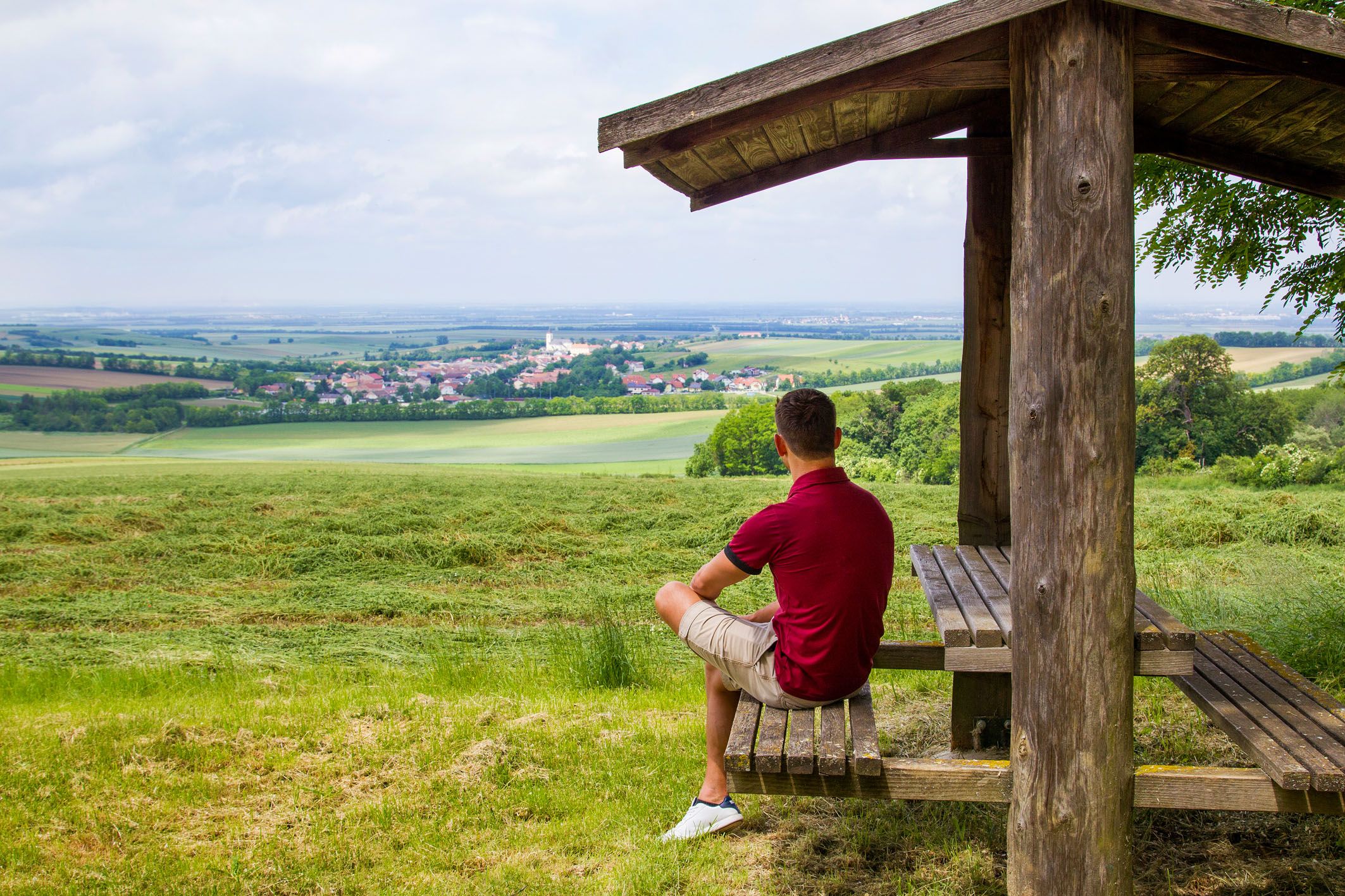 A man sits under a wooden shelter and looks out over a wide landscape with fields and a village in the distance.