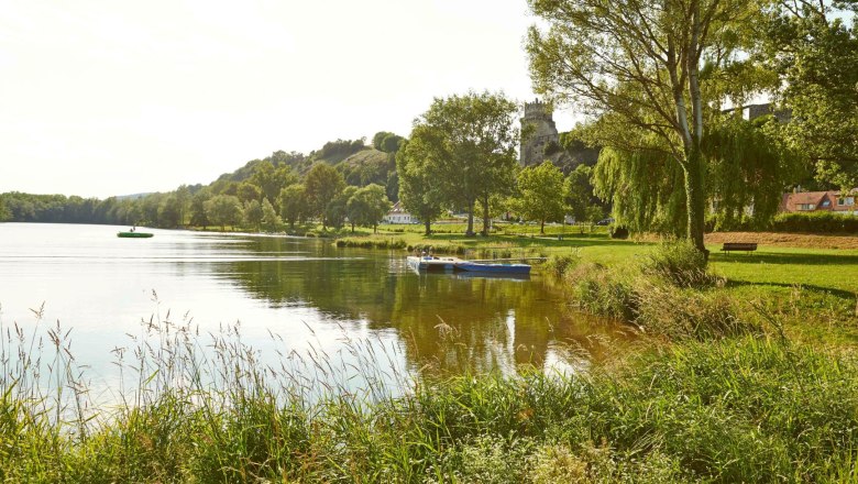 A calm lake with a boat, surrounded by green trees and meadows, with a castle on a hill in the background.
