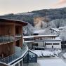 Winter landscape with snow-covered buildings and trees, in the foreground a modern wooden building with balconies.