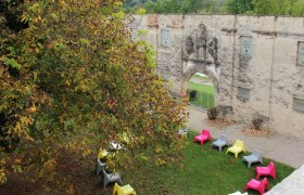 Colorful chairs in a circle on a meadow in front of an old stone wall with an archway.
