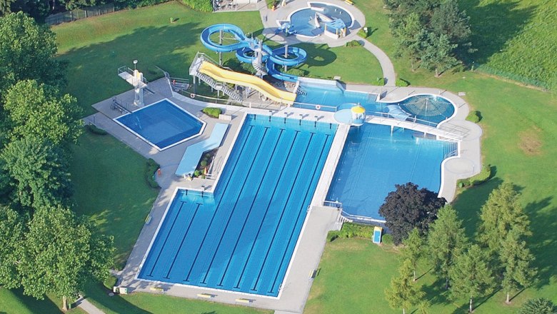 Aerial view of an outdoor pool with several pools and water slides, surrounded by green trees and lawns.