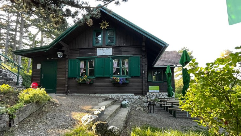A mountain hut with green shutters and flower boxes, surrounded by trees and garden furniture.