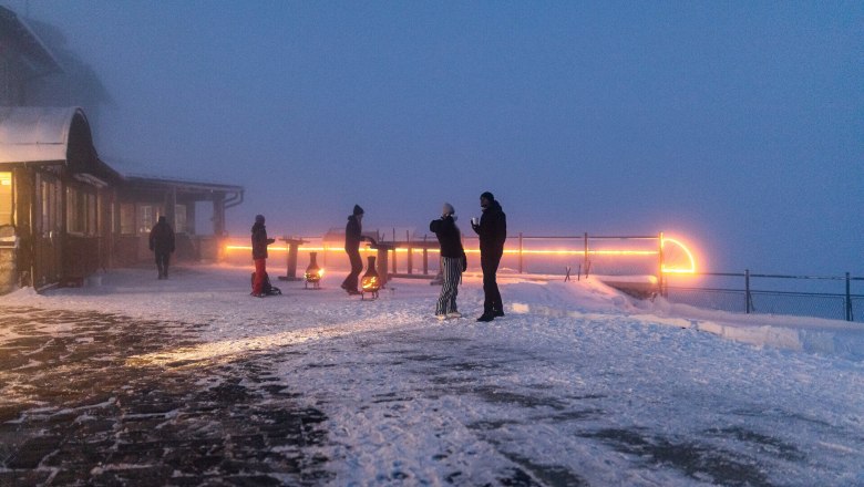 People standing on a snow-covered mountain terrace in the fog, illuminated by warm lights at dusk.