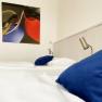 A guest room with a bed, blue cushions, reading lamps and an abstract painting on the wall.
