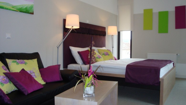 Premium bon-vivant rooms Zur Schonenburg, © Gasthof Hotel "Zur Schonenburg" Modernly furnished hotel room with double bed, sofa and decoration in shades of purple and green.