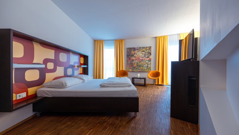Modern hotel room with double bed, colorful wall pattern, two orange chairs and yellow curtains.