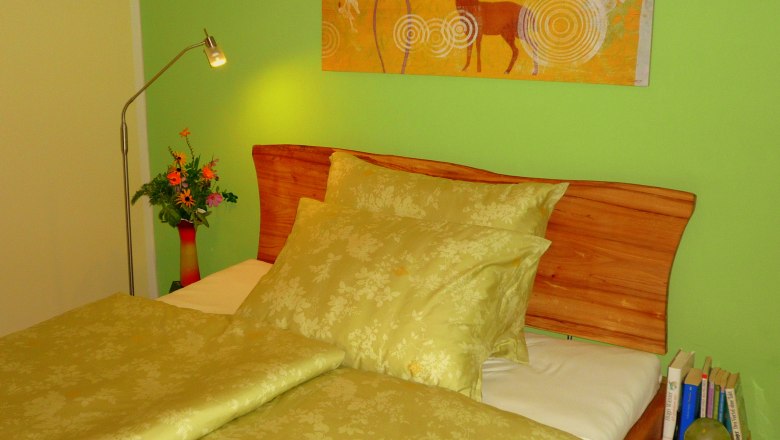 The small pension, © Die kleine Pension Cozy bedroom with green bedding, wooden headboard, green wall and deer picture. There is a lamp and flowers on the bedside table.
