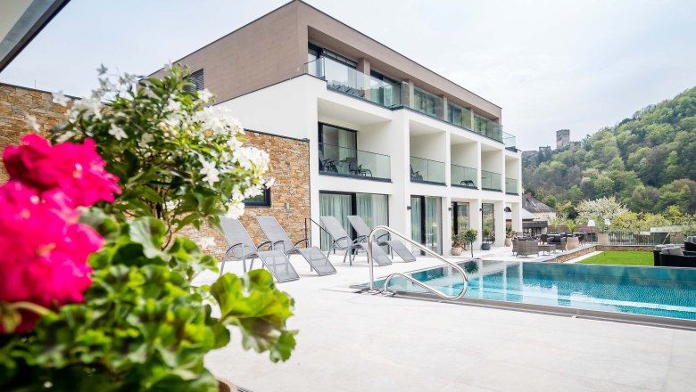 Modern building with pool and sun loungers, surrounded by green countryside and a castle in the background.