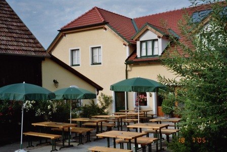A wine tavern garden with wooden benches and green parasols in front of a yellow building with red roof tiles.
