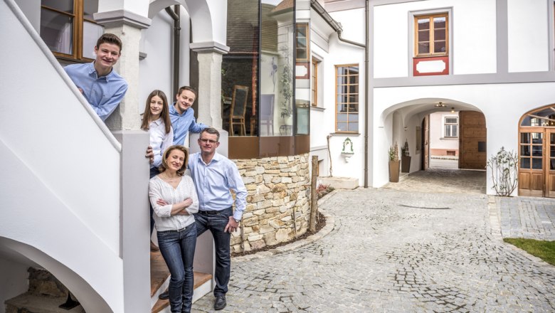 A smiling family stands on a staircase in front of a winery with a paved courtyard.