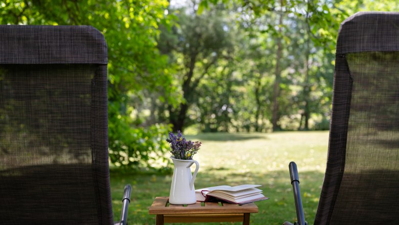 Two deckchairs in the garden with a table on which there is a vase of flowers and a book.