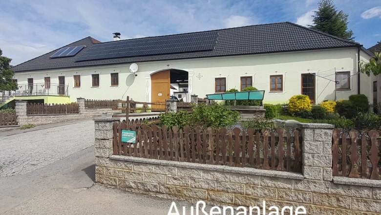 A large building with a yellow façade and black roof, surrounded by a garden and a wooden fence. The sky is blue with a few clouds.