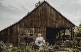 A cook stands in front of an old wooden hut with a bunch of herbs in his hand.
