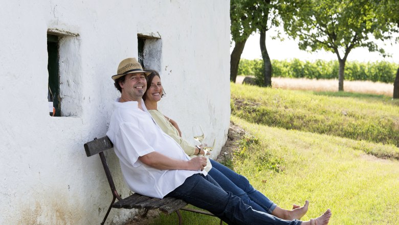 A couple sits relaxed on a bench against a white wall, wine glasses in hand, surrounded by a green landscape.