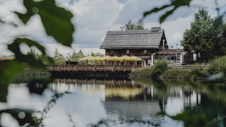 A rustic wooden house with yellow parasols is reflected in a calm lake surrounded by green nature.