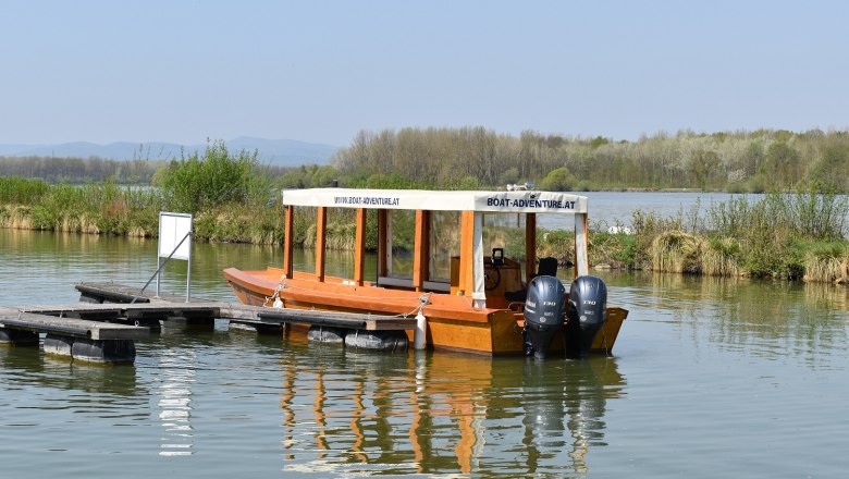 A wooden boat with two engines is moored on a jetty on a calm stretch of water, surrounded by green vegetation.