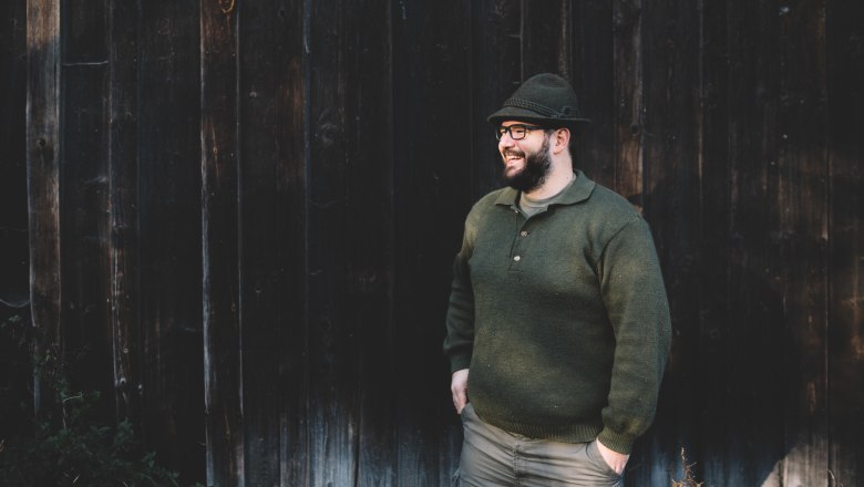 Man in green clothing and hat standing in front of a wooden wall.