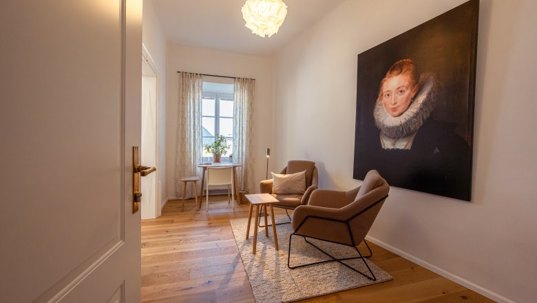 A cozy room with two armchairs, a table and a large portrait on the wall. A window with curtains lets in daylight.
