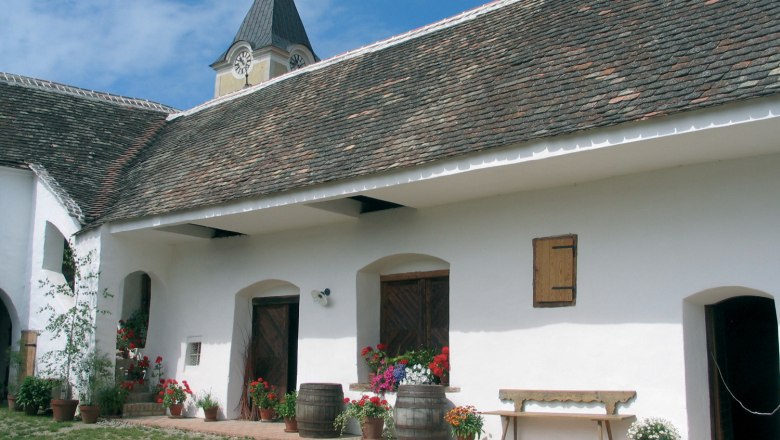 Traditional building with white façade, wooden doors and flowers in pots. A tower with a clock in the background.