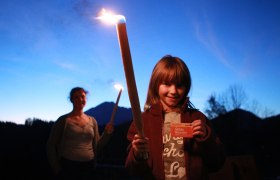 A child holds a burning torch and a map, in the background a woman with a torch, against a mountain backdrop at dusk.