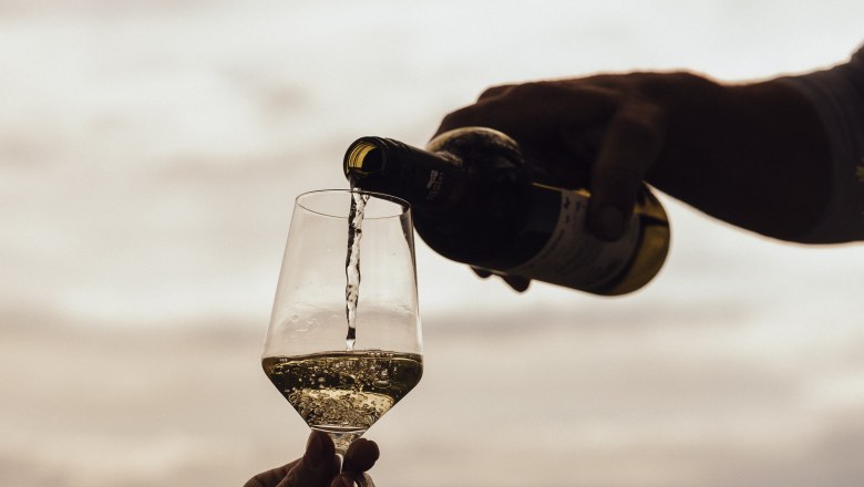 A hand pours white wine into a glass outside, with a cloudy sky in the background.