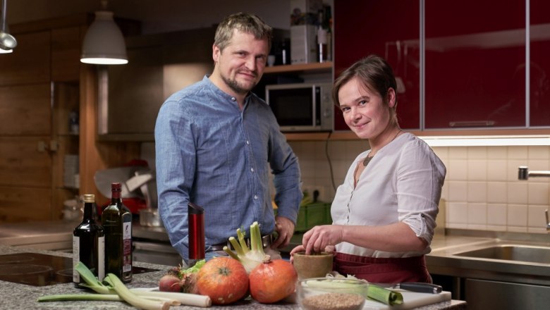 Two people in a kitchen with vegetables and bottles of wine on the worktop.